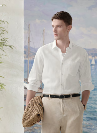 Man wearing a white button-up shirt and beige pants, holding a woven hat, standing near a scenic seaside backdrop with sailboats.