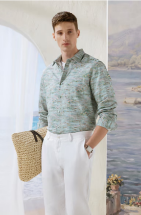 Man in a light green patterned long-sleeve shirt and white pants holding a woven straw bag, standing indoors near an arched window.