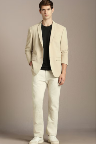 Man wearing a beige blazer over a black shirt, white pants, and white sneakers, standing against a neutral background with hands in pockets.