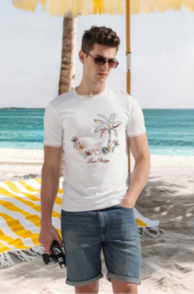 Man in white tropical print t-shirt and denim shorts standing on a sandy beach with ocean and palm tree in the background.