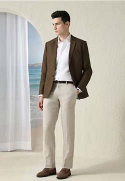 Man wearing a brown blazer, white shirt, beige pants, and brown loafers standing indoors near an arched window with a beach view.