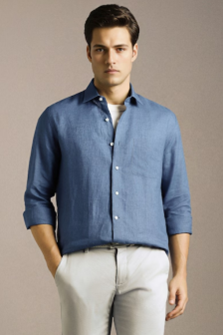 Man wearing a casual blue button-up shirt with rolled sleeves and light-colored pants, standing with hands in pockets against a plain background.