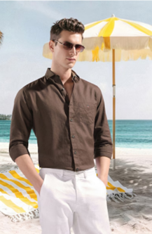 A man in sunglasses and a brown shirt stands on a sunny beach with white sand. A yellow-and-white striped umbrella and lounge chairs are in the background.
