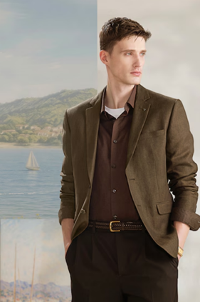 Man in brown blazer and dark shirt stands with hands in pockets against a backdrop of a lake and mountain landscape with a sailboat.