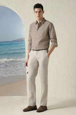 Man wearing a light brown button-up shirt and beige pants, standing by a white curved wall with a view of the ocean in the background.