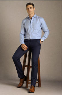 Man wearing a light blue dress shirt, dark blue pants, and brown shoes, sitting casually on a wooden stool against a plain background.