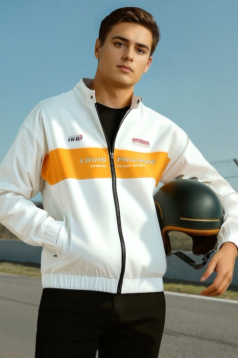 Young man in a white and orange Louis Philippe jacket holds a black helmet beside a racetrack.