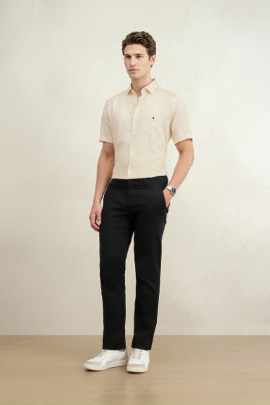 Full-length male model wearing a beige short-sleeve shirt, black trousers, and white sneakers, standing against a neutral studio backdrop.