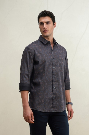 Man wearing a dark patterned button-up shirt and black pants, standing against a light textured wall.