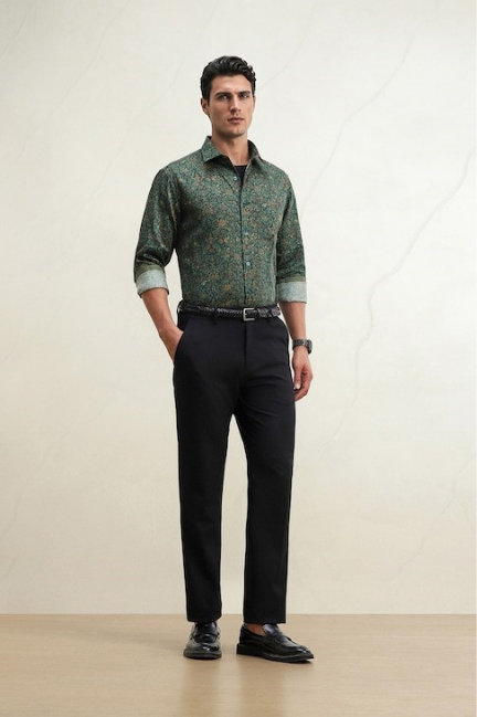 Man modeling a patterned green shirt, black trousers, belt, and dress shoes against a light wall.