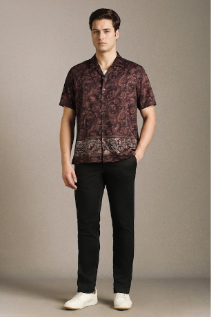 Young man modeling a paisley-patterned short-sleeve shirt, black pants, and white sneakers against a neutral studio background.
