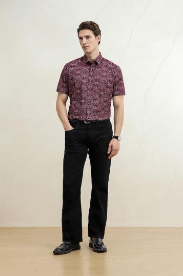 Young man modeling a burgundy patterned short-sleeve shirt, black trousers, and black shoes against a neutral studio background.